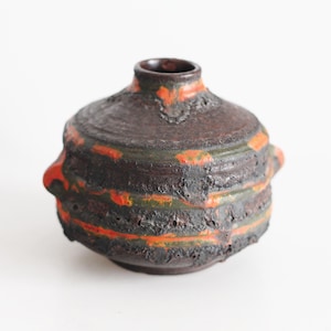 May include: A small, round ceramic vase with a dark brown, heavily textured, and rough surface. The vase features horizontal bands of bright orange and muted green glaze accents, a narrow neck, and two small, protruding handles on the sides.
