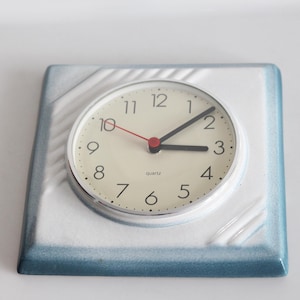 May include: A white ceramic clock with a blue border and a silver face. The clock has black numbers and hands, and a red second hand. The clock says "Quartz" at the bottom.