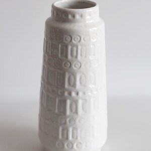 May include: A white ceramic vase with a textured surface. The vase has a cylindrical shape with a slightly flared top and a series of embossed rectangular and circular patterns. The vase is approximately 10 inches tall.