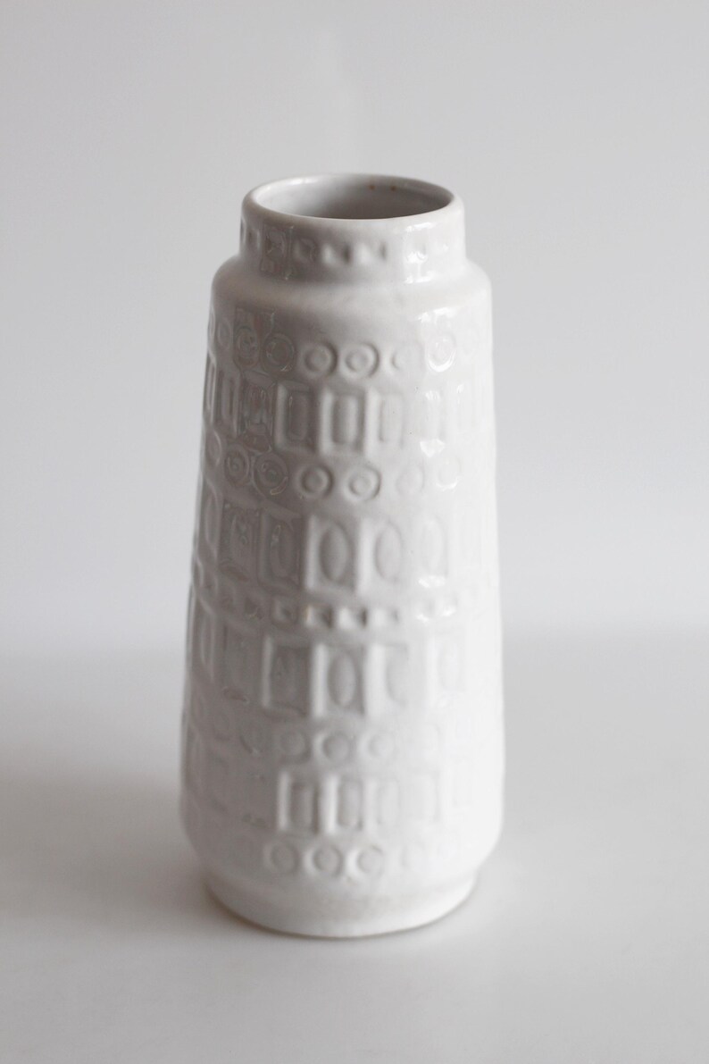May include: A white ceramic vase with a textured surface. The vase has a cylindrical shape with a slightly flared top and a series of embossed rectangular and circular patterns. The vase is approximately 10 inches tall.