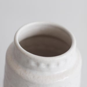 May include: A white ceramic vase with a cylindrical body and a slightly flared rim. The vase has a textured surface and a neutral color palette, suitable for various home decor styles. The opening is approximately 3 inches in diameter.
