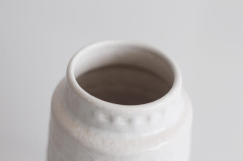 May include: A white ceramic vase with a cylindrical body and a slightly flared rim. The vase has a textured surface and a neutral color palette, suitable for various home decor styles. The opening is approximately 3 inches in diameter.