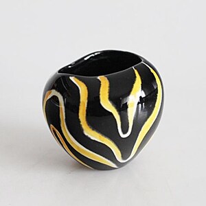 May include: A small, black ceramic vase with a glossy finish. The vase features a pattern of yellow and white curved lines. The opening of the vase is oval-shaped. The vase is a decorative item.