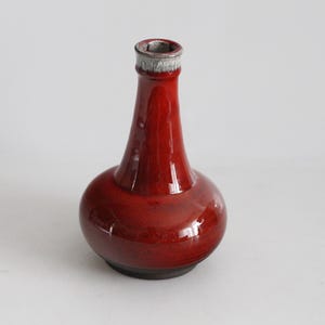 May include: A glossy, red ceramic vase with a bulbous base and a slender neck. The rim is silver. The vase is approximately 8 inches tall and has a dark base. The design is simple and elegant.