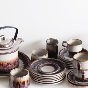 Puede incluir: Un juego de té de cerámica con tetera, tazas, platillos y una jarra de crema. El juego presenta una combinación de colores gris y marrón con un asa oscura en la tetera. Las tazas están apiladas y los platillos están ordenados.