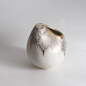 May include: A white ceramic vase with a unique shape and a gold-colored rim. The vase has a textured, gray and gold pattern on the upper half, with a smooth white base. The design gives it a vintage or antique feel.