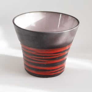 May include: A black and red ceramic cup with a white interior. The cup has a wide mouth and a narrow base. It features a striped pattern of red and black lines.