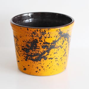May include: A ceramic planter with a bright yellow exterior and a black interior. The yellow surface is decorated with black splatter paint. The planter has a cylindrical shape and a wide opening, suitable for various plants.
