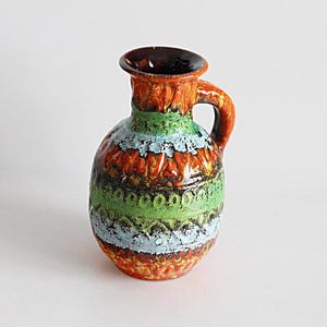 Puede incluir: Jarra pequeña de cerámica con asa. La jarra presenta un diseño texturizado con bandas naranjas, verdes y azules. El borde y el interior son de color marrón oscuro. La jarra es probablemente vintage o antigua.