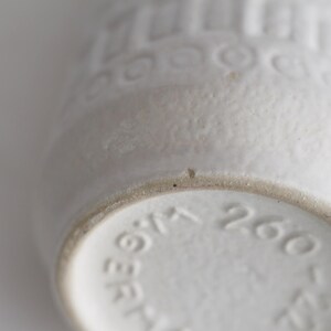 May include: Close-up of a white ceramic vase with a textured surface and embossed patterns. The base of the vase has stamped numbers and text. The vase is a neutral color and has a vintage aesthetic.