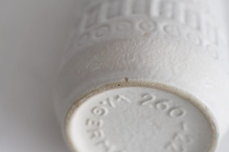 May include: Close-up of a white ceramic vase with a textured surface and embossed patterns. The base of the vase has stamped numbers and text. The vase is a neutral color and has a vintage aesthetic.