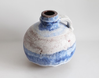 May include: A round ceramic jug with a handle, featuring a color palette of white, blue, and brown. The jug has a dark brown opening and a white handle. The design includes horizontal bands of color, creating a textured appearance.