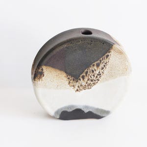 Puede incluir: Un jarrón de cerámica con forma ovalada redondeada. El jarrón está decorado con un esmalte marrón, blanco y negro, creando un efecto de capas. El jarrón tiene una pequeña abertura en la parte superior para flores.