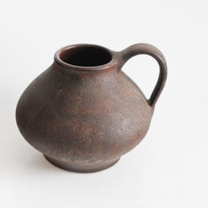 Puede incluir: Jarra de cerámica marrón con superficie texturizada, forma redondeada y asa curva. La jarra tiene un interior oscuro y una pequeña base circular. Diseño sencillo y rústico.