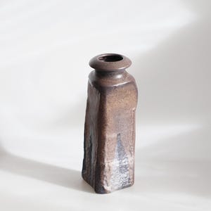 May include: A tall, brown ceramic vase with a rectangular body and a rounded top. The vase has a textured surface and a dark brown color with lighter brown and black accents. The vase is approximately 8 inches tall.