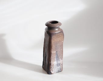 May include: A tall, brown ceramic vase with a rectangular body and a rounded top. The vase has a textured surface and a dark brown colour with lighter brown and black accents. The vase is approximately 20 cm tall.