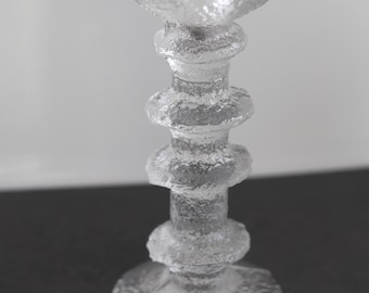 May include: A clear glass candle holder featuring a textured, ice-like surface. The design includes a wide circular base, a central stem with three distinct horizontal ring-shaped protrusions, and a wide, flat top with a central opening for a candle.