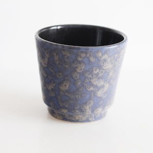 Op de afbeelding: Een keramische bloempot met een gespikkelde blauwe en bruine glazuur. De binnenkant is zwart. De bloempot heeft een licht uitlopende vorm en een kleine, verhoogde basis. De achtergrond is wit.