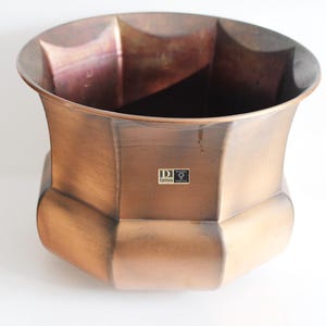 Op de afbeelding: Een grote, achthoekige koperen plantenbak met een licht verweerde afwerking. De plantenbak heeft een label op de bodem met de tekst "DD Designs".
