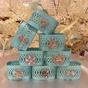 May include: A set of eight teal blue napkin rings with decorative cutouts and jeweled embellishments. The rings are stacked in a pyramid shape.