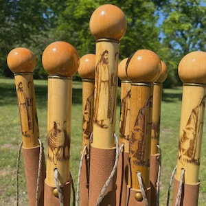 May include: A collection of wooden walking sticks featuring a smooth spherical top, a cylindrical shaft with pyrography illustrations of a shepherd and sheep, and a tan leather grip section secured with brass rivets and a thin cord lanyard.
