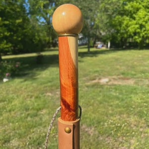 May include: A wooden walking stick with a light brown, spherical handle. The shaft is a mix of reddish-brown and light wood tones. A leather strap with a brass button and a patterned cord is attached near the bottom.