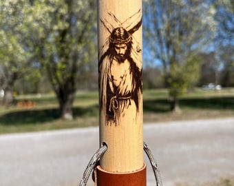 May include: A cylindrical wooden walking stick featuring a dark wood-burned illustration of a figure wearing a crown of thorns and carrying a cross. The stick includes a brown leather grip section and a grey speckled cord threaded through holes.
