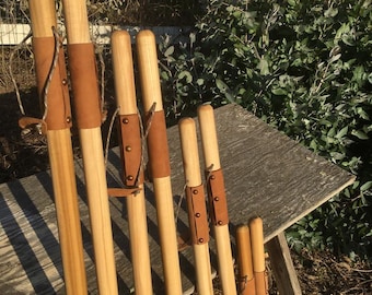 May include: A set of eight wooden walking sticks of varying lengths, each featuring a smooth, light-colored cylindrical shaft and a tan leather grip secured with metal rivets and a thin leather cord loop attached near the top handle area.