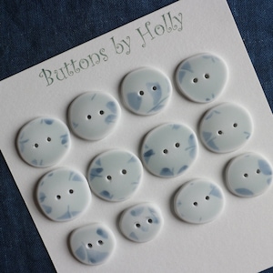 Könnte beinhalten: Ein Set mit 12 blauen und weißen Keramikknöpfen mit einem marmorierten Design. Die Knöpfe sind in einem Raster auf einer weißen Karte mit dem Text "Buttons by Holly" angeordnet.