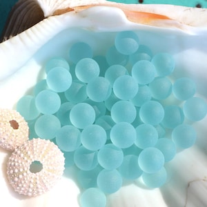 Puede incluir: Una colección de cuentas de vidrio esmerilado en color azul claro. Las cuentas son redondas y lisas, y están dispuestas en una pila en una concha de mar.
