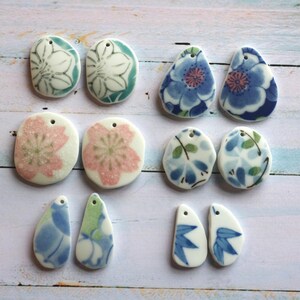 May include: A collection of sixteen ceramic pendants arranged in pairs, featuring various floral and botanical patterns in shades of blue, pink, green, and white. Each piece has a small hole at the top for stringing, with diverse shapes.