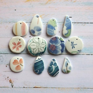 Puede incluir: Una colección de once colgantes de cerámica de diversas formas y tamaños, cada uno con diseños pintados a mano que incluyen motivos florales, patrones abstractos en azul y verde, una libélula y hojas de trébol rosa suave, todos con un pequeño orificio.