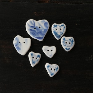 Puede incluir: Un conjunto de siete botones de cerámica en forma de corazón con patrones florales azules y blancos. Los botones son de diferentes tamaños y tienen dos agujeros cada uno.