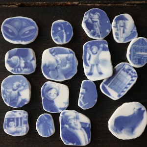 May include: A collection of 15 blue and white ceramic tiles featuring various images, including a snowman, a house, a person in a winter coat, and a dog. The tiles are all different shapes and sizes.