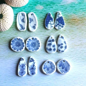 May include: A collection of ceramic earrings with blue and white floral and abstract designs. The earrings are in various shapes, including teardrops and circles. The earrings are displayed on a light blue and green background.