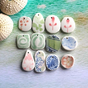Puede incluir: Una colección de colgantes de cerámica de varias formas, incluyendo ovalados, de lágrima y redondos. Cada colgante es blanco con diseños florales o abstractos pintados a mano en verde, rosa y azul. Los colgantes tienen pequeños agujeros para ensartar.