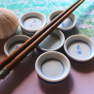 Puede incluir: Un conjunto de pequeños cuencos de sake de cerámica blanca con bordes azules y caracteres japoneses, dispuestos alrededor de un par de palillos de madera. También hay una concha de erizo de mar texturizada y de color claro. El fondo tiene un efecto de acuarela.