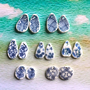 May include: A selection of white ceramic pendants with blue patterns. The pendants are in various shapes, including teardrops and circles, and feature floral, geometric, and abstract designs. Each pendant has a small hole for stringing.
