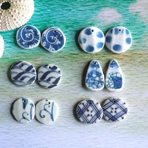 May include: A collection of white ceramic beads with blue patterns, including dots, swirls, and geometric designs. The beads are in various shapes, such as circles and teardrops, and have small holes for stringing. The background is a watercolor-style wash of green and blue.