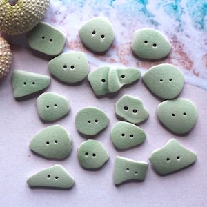 May include: A collection of seafoam green ceramic buttons in various organic shapes, each with two holes for sewing. The buttons have a crackle glaze finish and are scattered on a light pink and blue surface, with sea urchin shells in the background.