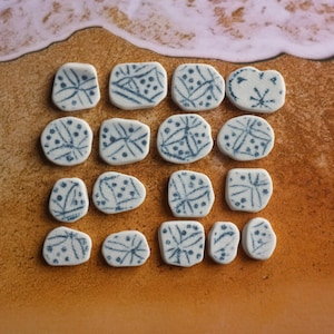 Puede incluir: Una colección de pequeñas baldosas de cerámica blancas con diseños abstractos azules. Las baldosas, de varias formas, están dispuestas sobre una superficie arenosa con una ola en el fondo.