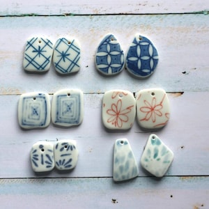 May include: A collection of twelve ceramic pendants arranged in six pairs. The pendants feature various hand-painted patterns in blue and orange, including geometric grids, floral motifs, and abstract shapes, each with a small hole for hanging.