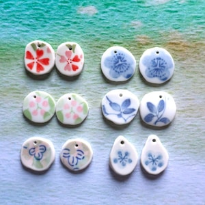 May include: A collection of small, white ceramic pendants with floral designs in red, pink, blue, and green. Each pendant has a small hole for stringing. The pendants are in various shapes, including round and teardrop.