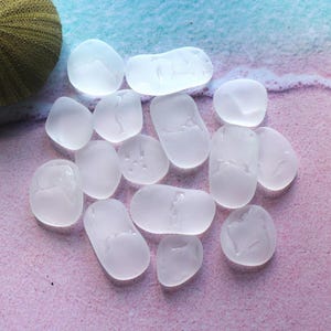 Op de afbeelding: Een verzameling van vijftien gladde, matwitte stukjes zeeglas in verschillende vormen en maten. Elk stuk heeft een matte, doorschijnende afwerking met afgeronde randen en is losjes over het beeld verspreid zonder specifiek patroon.