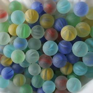 Pode incluir: Uma coleção de bolinhas de vidro coloridas em vários tons de azul, verde, vermelho e amarelo. As bolinhas são lisas e têm um acabamento fosco.