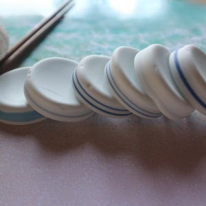 Puede incluir: Una colección de posavasos de cerámica blanca con rayas azules. Los posavasos son redondos y tienen un tema náutico. Una concha y palillos están en el fondo.