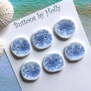 May include: Six white ceramic buttons with blue floral designs and two holes each. The buttons are arranged on a white card with the text "Buttons by Holly" in a script font. The background includes a seashell.