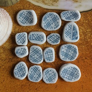 May include: A collection of 14 white ceramic tiles with a blue criss-cross pattern. The tiles are arranged in a grid pattern on a brown surface.