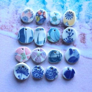 May include: A collection of small, ceramic pendants with various floral and abstract designs. The pendants are primarily white with blue, pink, and yellow accents. Each pendant has a small hole for stringing. The overall theme is handmade jewelry.