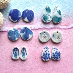 Puede incluir: Una colección de colgantes de cerámica de varias formas, con diseños florales y abstractos en azul y blanco. Los colgantes tienen pequeños agujeros para ensartar y se exhiben sobre una superficie rosa.